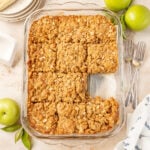 Apple pie bars in a glass 9 x 13 baking pan on a stone surface with forks plates and apple surrounding it.