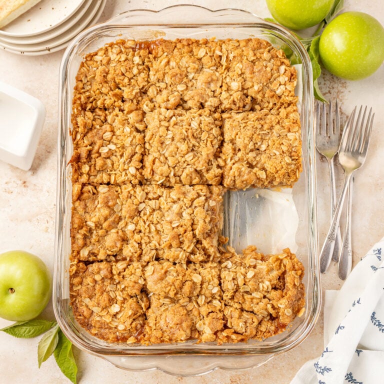 Apple pie bars in a glass 9 x 13 baking pan on a stone surface with forks plates and apple surrounding it.