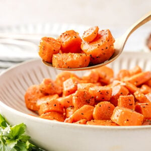 Close-up of Brown Sugar Glazed Carrots on a spoon above a serving bowl, coated in a glossy butter and brown sugar glaze.