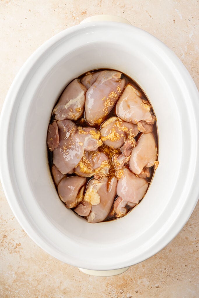 Raw boneless chicken thighs coated in teriyaki sauce in a white slow cooker before cooking.