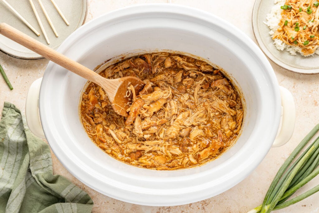 Shredded teriyaki chicken mixed with sauce inside a white slow cooker with a wooden spoon.