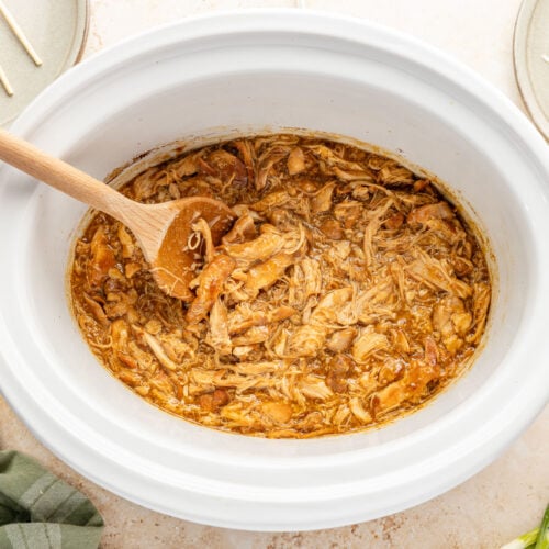 Shredded teriyaki chicken mixed with sauce inside a white slow cooker with a wooden spoon.